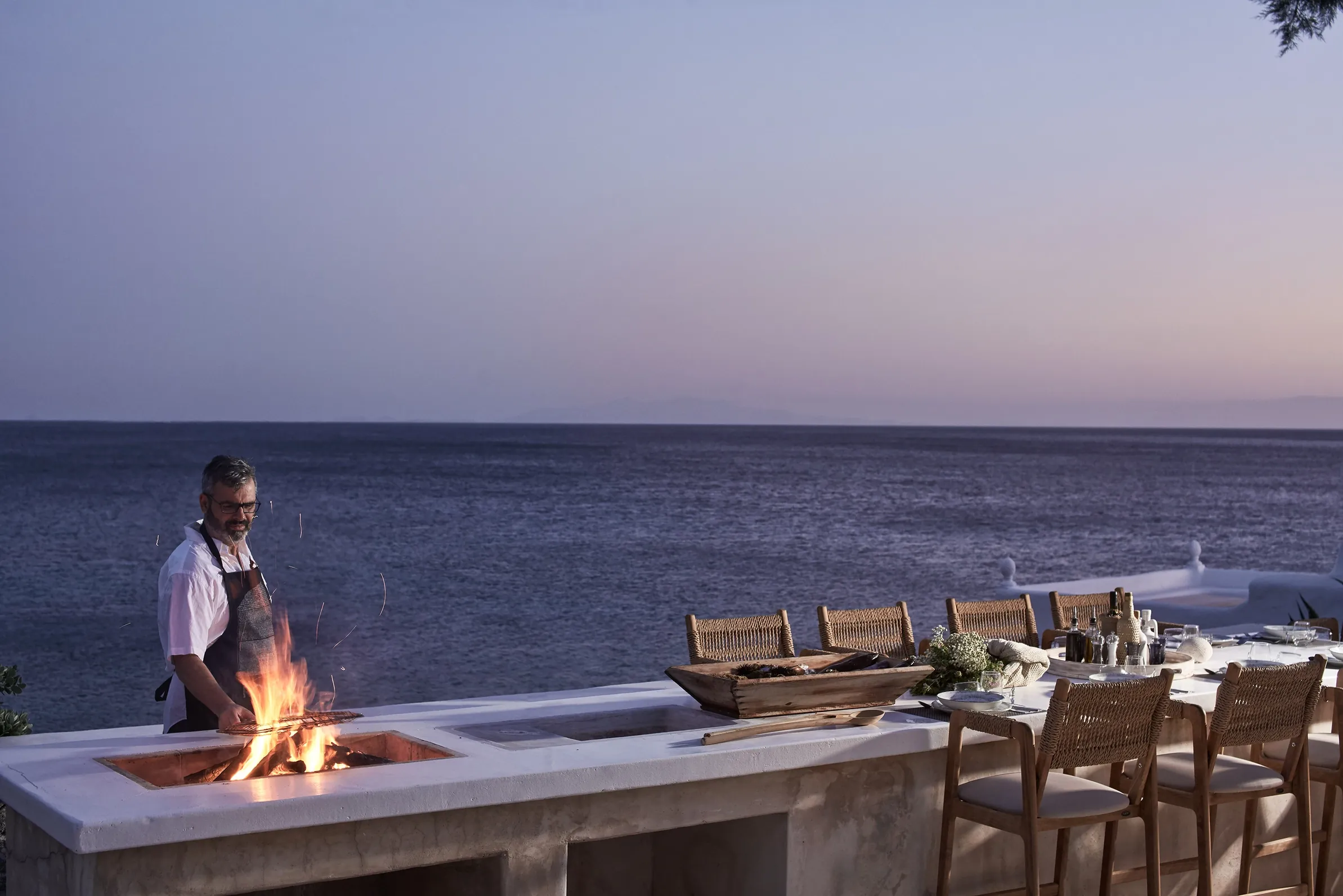 Chef grilling over an open fire beside the long dining table on the sea-view terrace at dusk