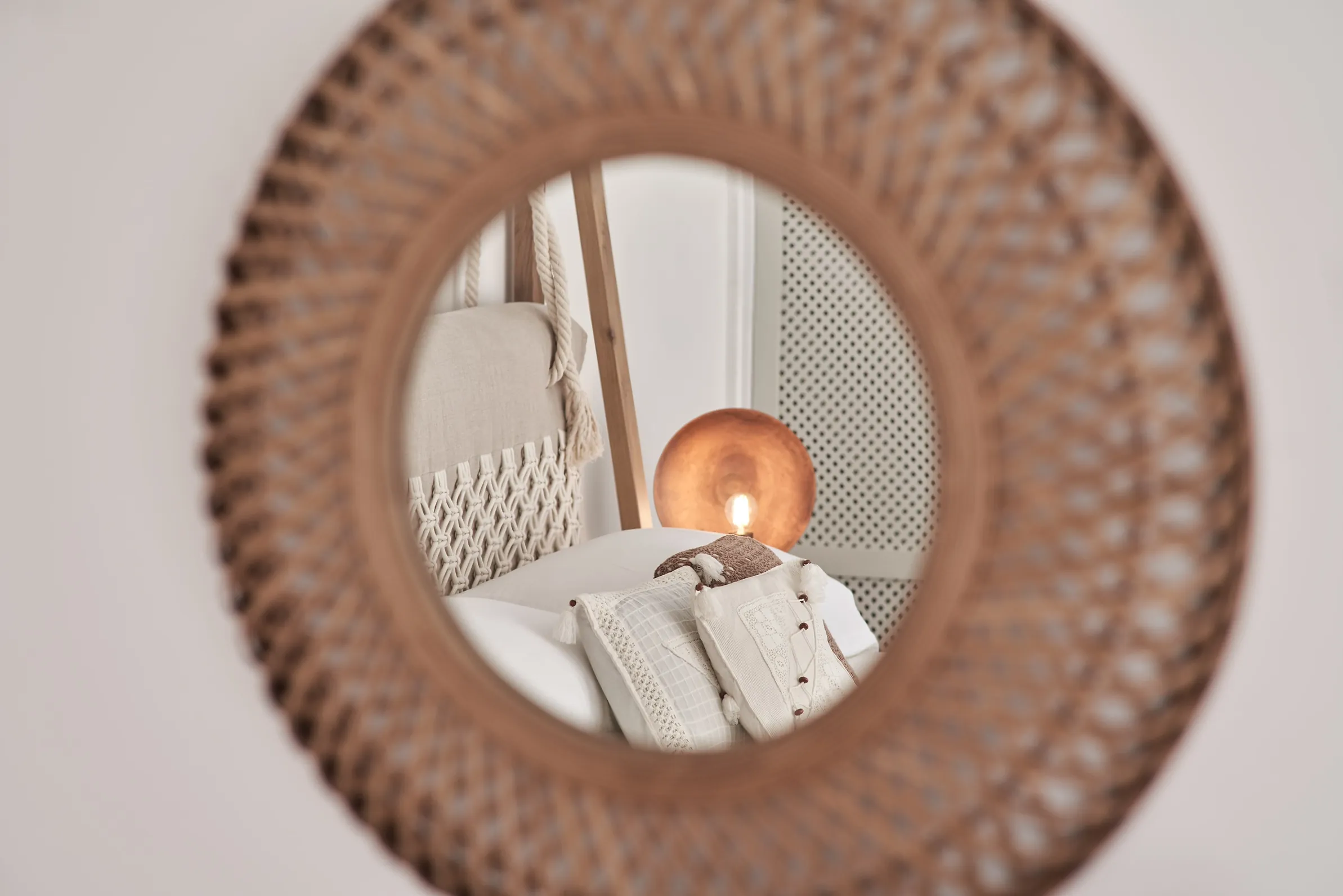 Rattan-framed round mirror reflecting a bedroom's cushions and lamp