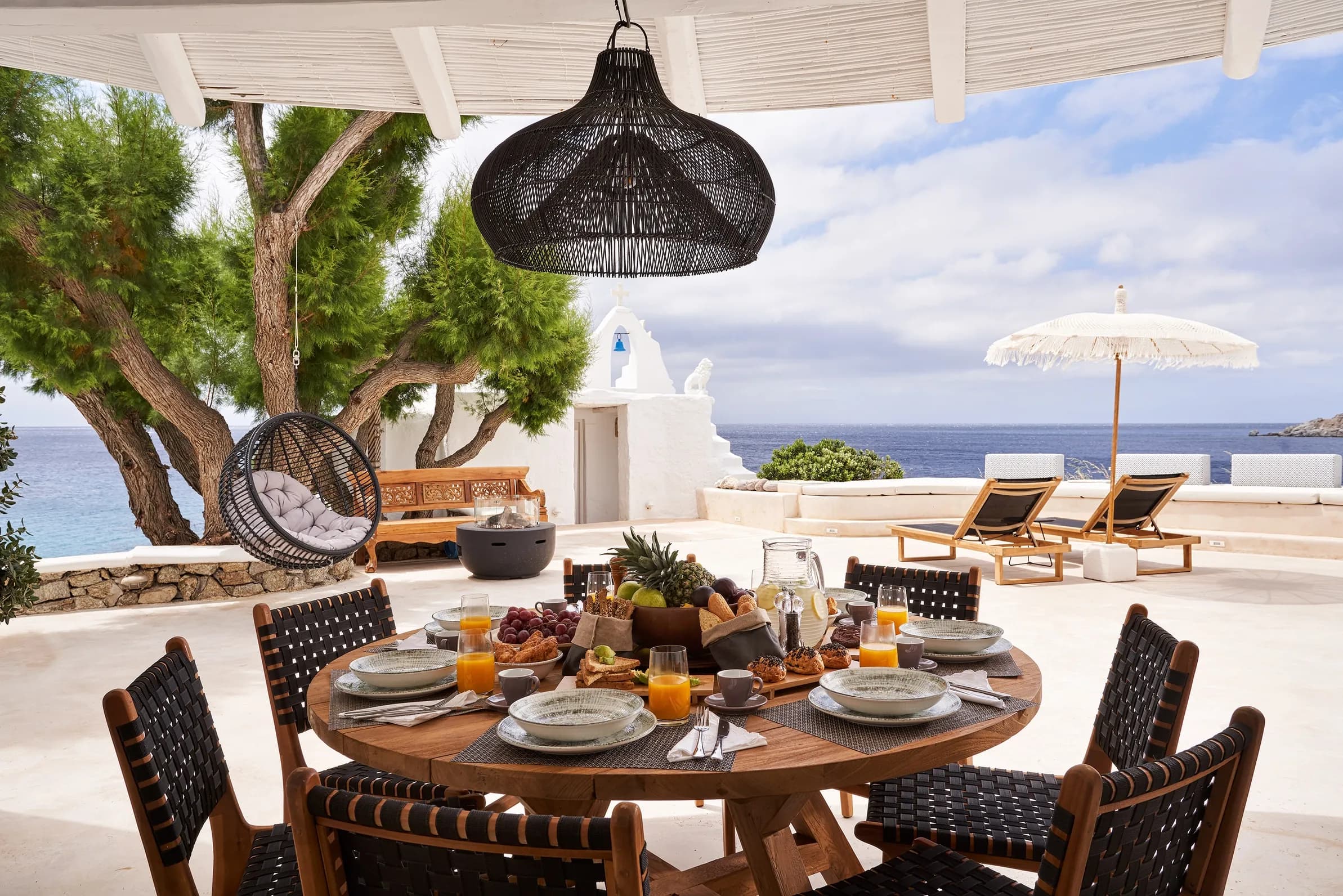 Breakfast laid on a round table under the pergola, with sun loungers, a parasol and the chapel beyond