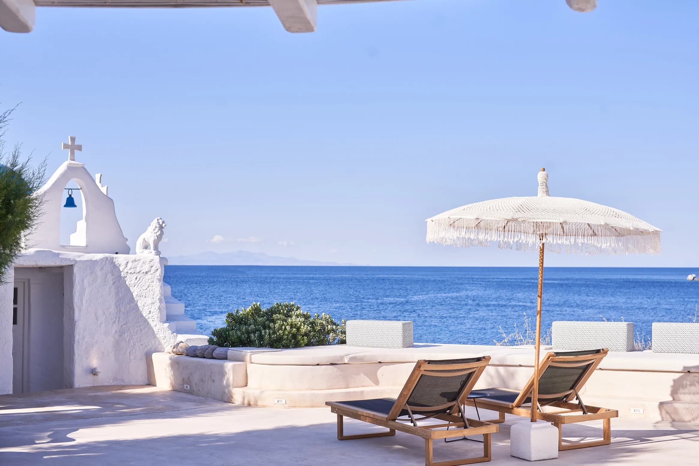 Sun terrace at Santa Katerina Mykonos with loungers, a fringed parasol and the whitewashed chapel above the Aegean Sea